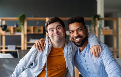 Young man with Down syndrome and his tutor with arms around looking at camera indoors at school